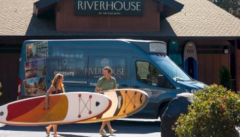 Two people carrying stand up paddleboards next to Riverhouse Resort's airport and area shuttle on a sunny day.