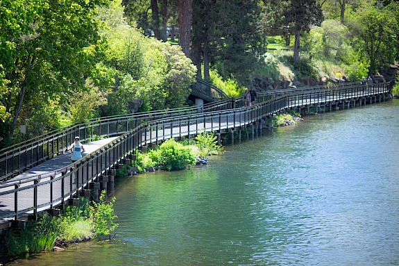 A person jogs on a winding wooden path beside a calm river, surrounded by lush green trees and foliage in Bend Oregon