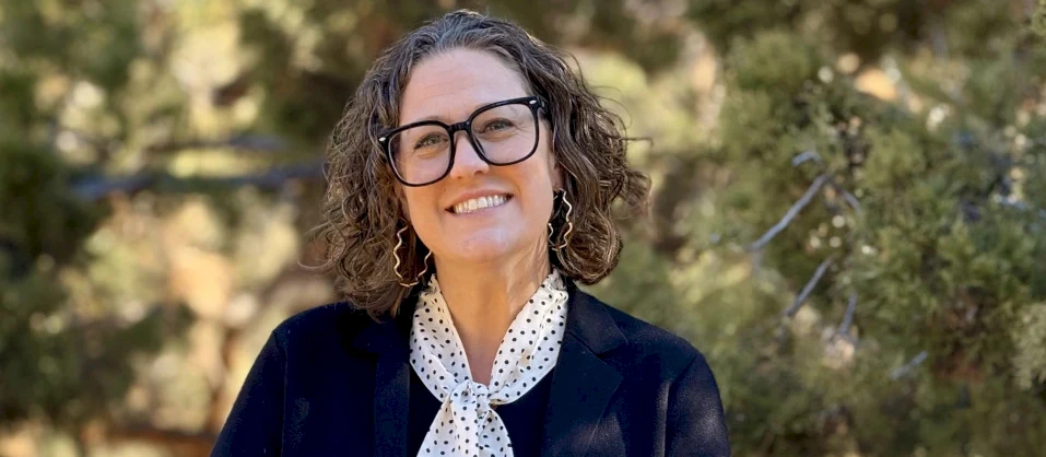 A woman with shoulder-length curly hair and glasses smiles outdoors, wearing a dark blazer and white polka-dot scarf, in a leafy setting.