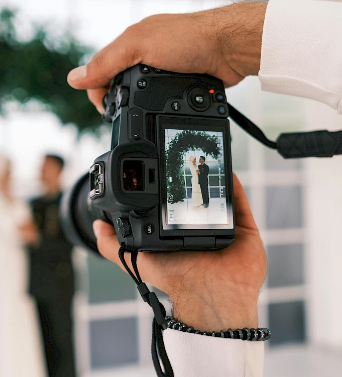 A photographer holding a camera takes a picture of a couple in the background, with the image on the LCD showing the scene.