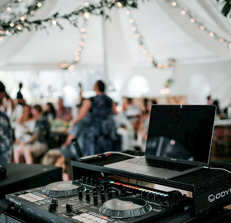 A DJ setup with a laptop and mixing console inside a decorated event tent, guests dancing in the background, twinkling lights overhead.